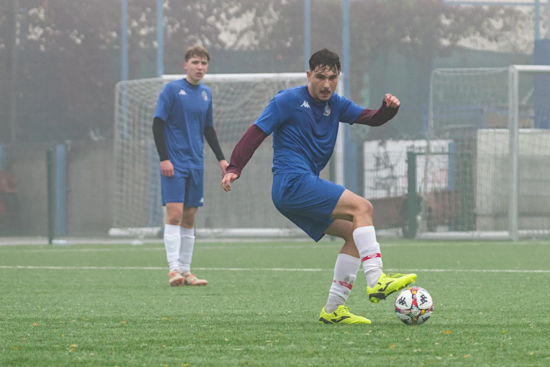 Dva nogometaša u plavim uniformama na zelenom terenu, jedan kontrolira loptu jarko žutim kopačkama.